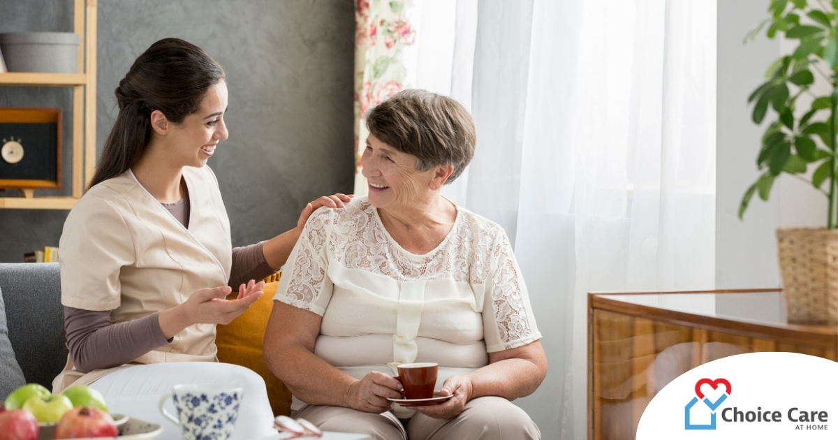 Smiling caregiver with her hand on an older woman’s shoulder as they share tea on the couch, showing home care vs assisted living